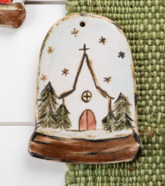 A hand painted pottery ornament depicting a church design with a snowy scene and Christmas trees.