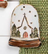 A hand painted pottery ornament depicting a church design with a snowy scene and Christmas trees.