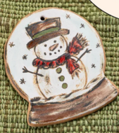 Hand-painted ceramic holiday ornament featuring a snowman design with a red scarf and a black hat, set against a white background with snow.